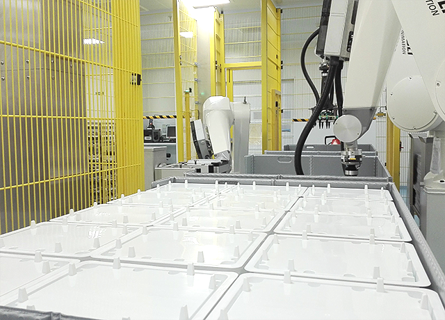 A picture of a pharmaceutical production line showing 3 pallet size plastic containers loaded with stacked tubs separated with plastic lids. 3 robots equipped with grippers handling tubs with lids and trays are visible in the background.