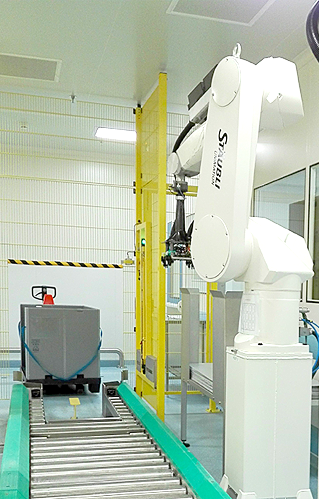 A picture of a pharmaceutical production line showing an empty stainless-steel roller conveyor with a pallet size container and 2 robots equipped with grippers in the background.