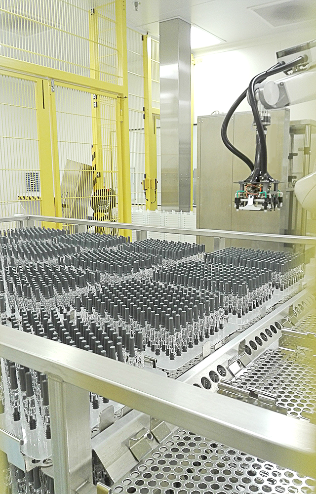 A picture of a pharmaceutical sterilization production line showing a cart with several trays turned upside down with nested syringes securely held in place and a robot equipped with a gripper in the background